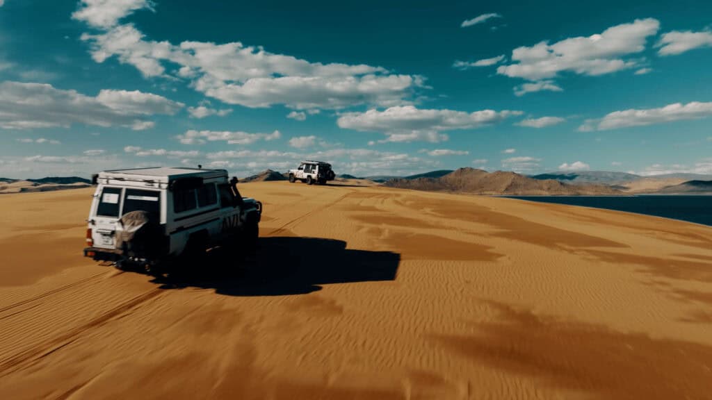Two offroad cars in Mongolia reaching the top a sand dune with a brilliant blue sky above and amazing scenery in the distance