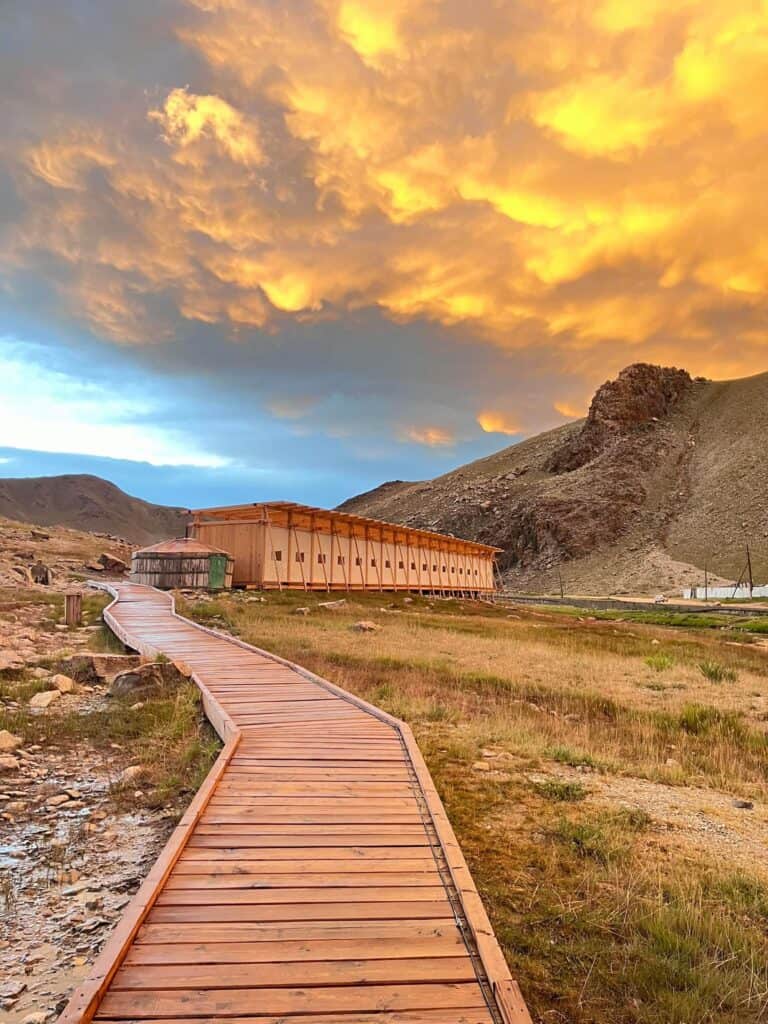 ممر خشبي يؤدي إلى نبع ساخن محاط بتضاريس صخرية وسماء نابضة بالحياة.