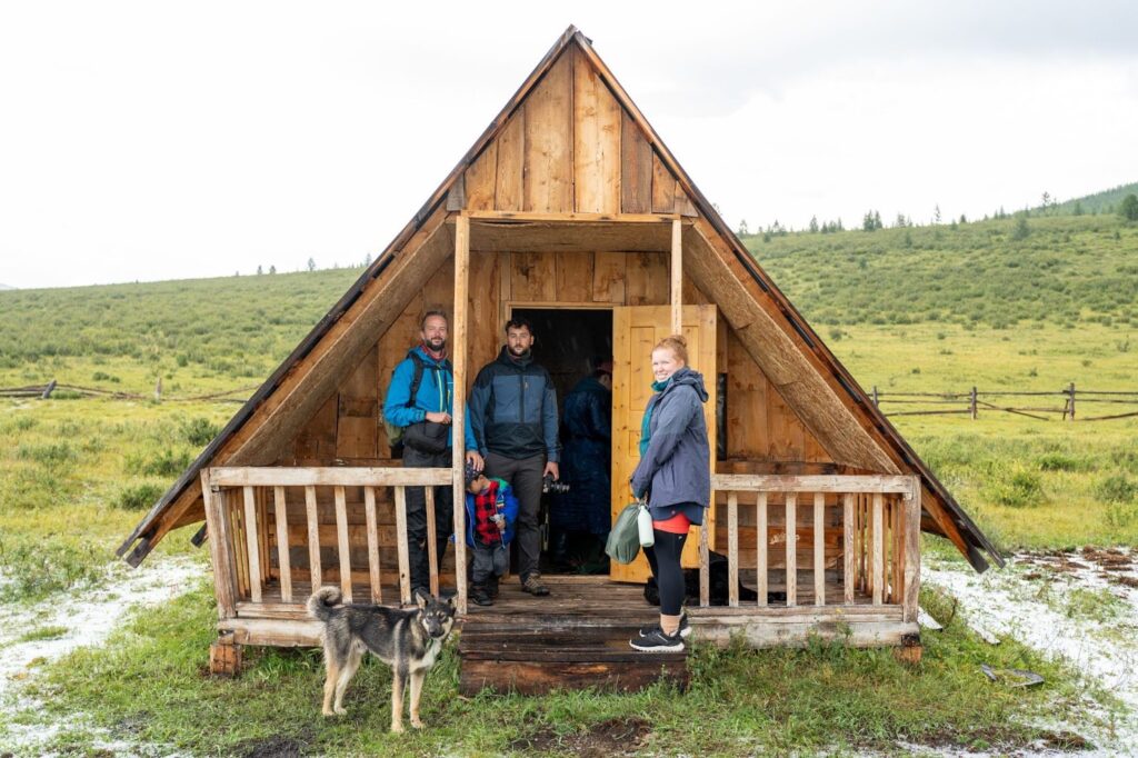 Three friends and a dog inside the doorway of a triangular cabin with some snow scattered around.