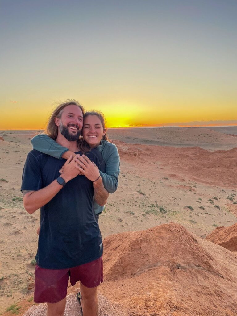 Couple on a Mongolia road trip pose in front of the sunset in the pink and orange desert.