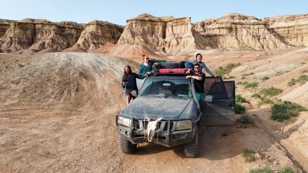 Two couples hanging out of the doors of a 4x4 vehicle with a cow skull on the front. Behind them the desert cliffs in Mongolia.