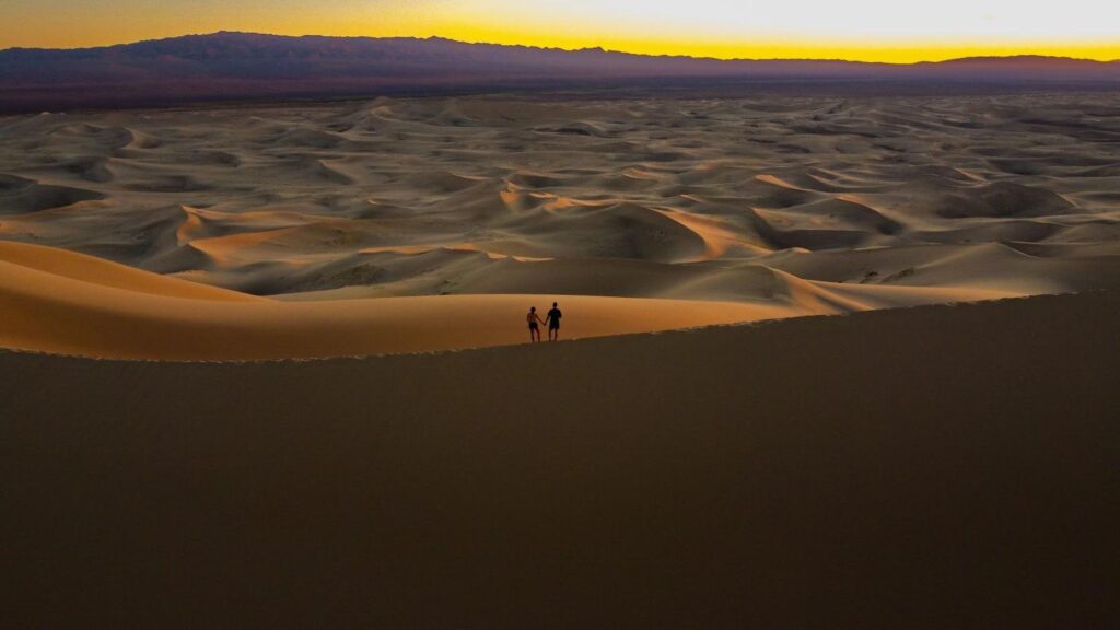 A couple stands on the top of a massive sand dune at sunset.