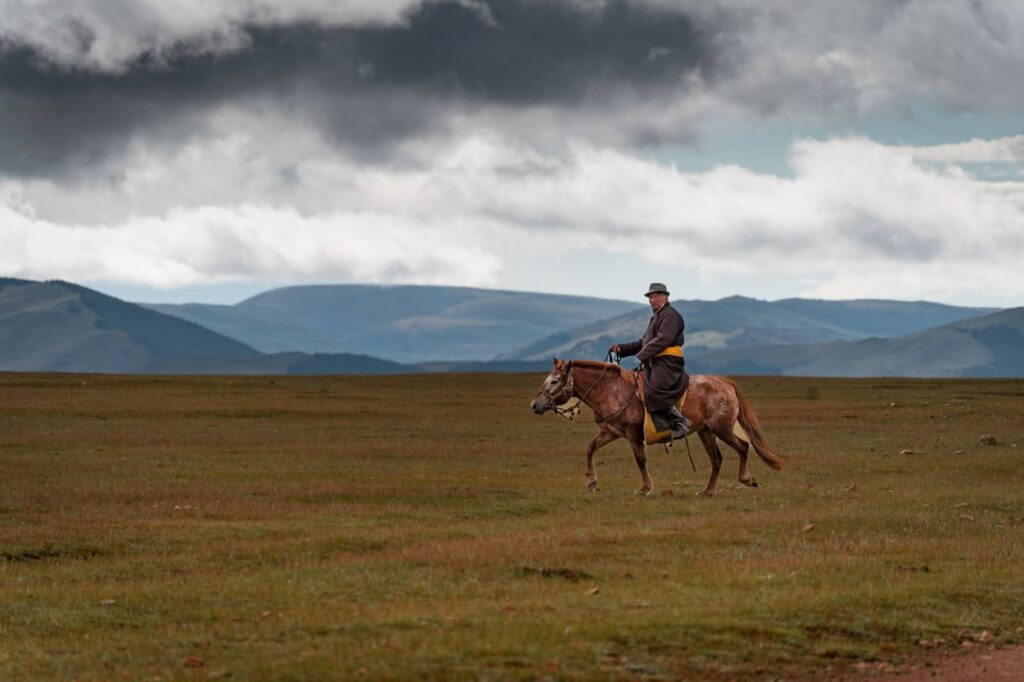 A mongolian man riding a horse through the steppe.